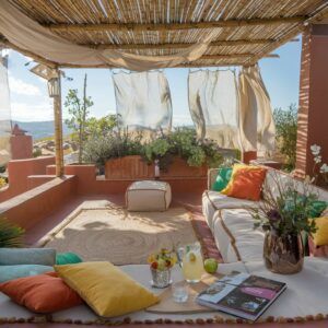 a small outdoor space with a earthy tones. There's a pergola with billowy fabric strung across it. Under the pergola, there's a square trimmed jute rug. There's a big soft sofa with color scatter cushions placed on the sofa.There are low walls and beautiful views in the background and it like an inviting space to relax.